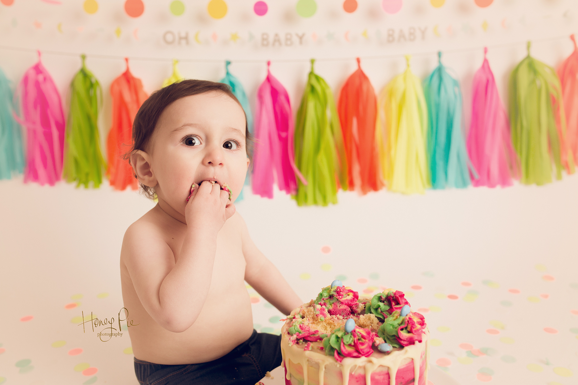 Baby getting a handful of cake in his mouth at photography session surrounded by neon party decorations