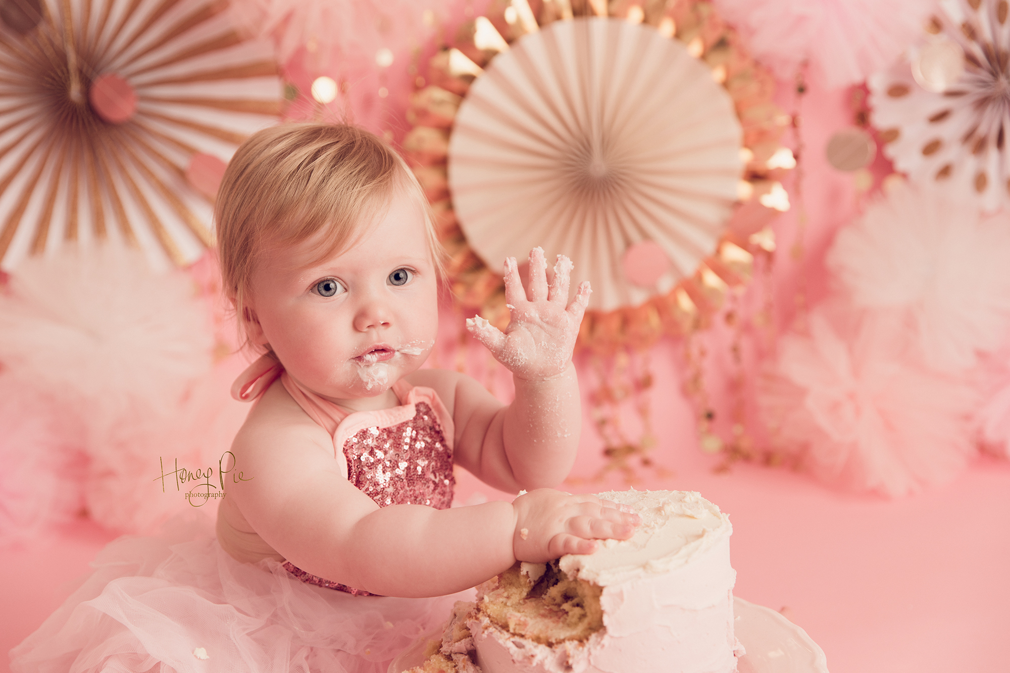 Baby girl waving to camera with cake all around her mouth & hands during her cake smash photography session in Worthing