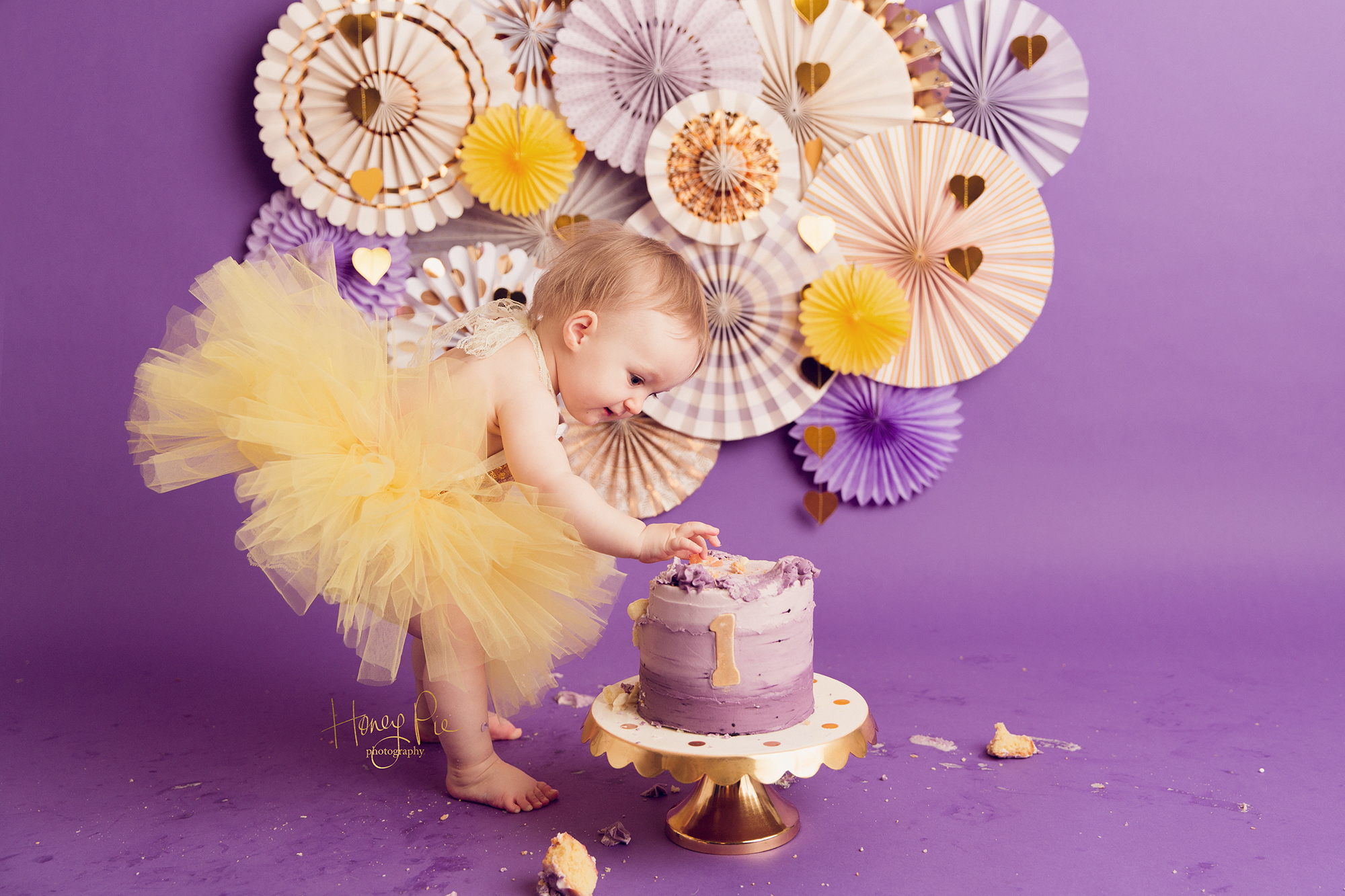 Baby delicately picking the icing off her first birthday cake wearing a bright yellow tutu