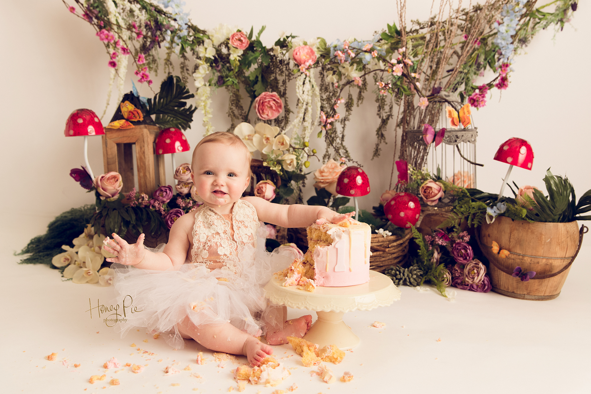 Baby in a fairy garden with toadstools, flowers & lanterns smashing her first birthday cake