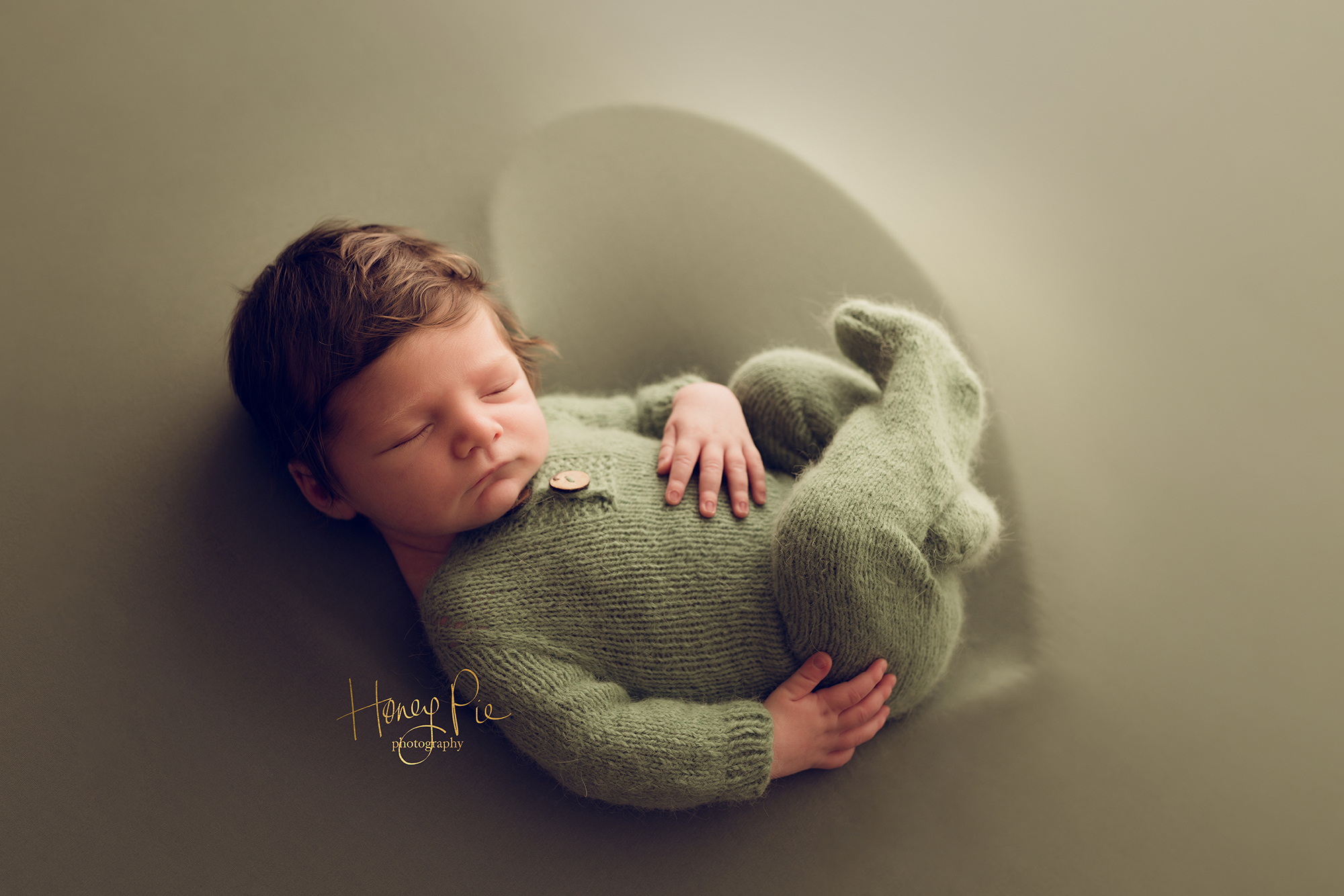 Newborn baby boy dressed in green, with lots of dark hair sleeping curled up in a heart