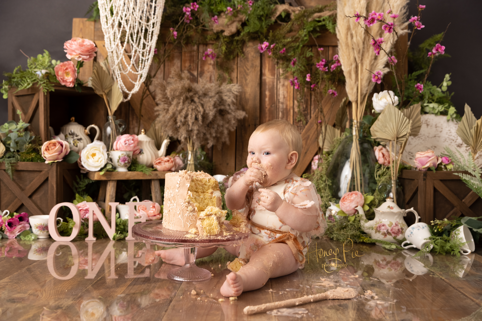 Baby girl chowing down on a chunk of birthday cake with vintage tea cups & pampas grass decor surrounding her